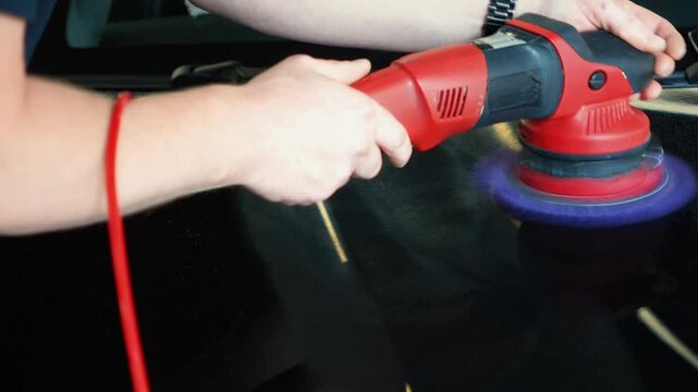 Close up of a professional car detailing process. Mans hands hold a red electric orbital dual action polisher machine with a purple microfiber wool pad buffer tool to polish a black auto paint hood.