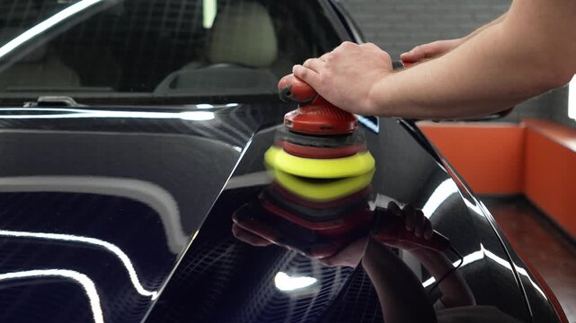 Professional car detailer worker polishes a luxury vehicle hood paint using an electric dual action orbital polishing machine tool with a yellow foam pad buffer in a garage workshop service.