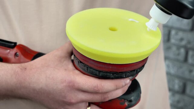 Close up of a professional car detailer applying white polishing compound paste drops on a yellow foam pad buffer tool of an electric orbital dual action polisher machine in a vehicle detailing garage