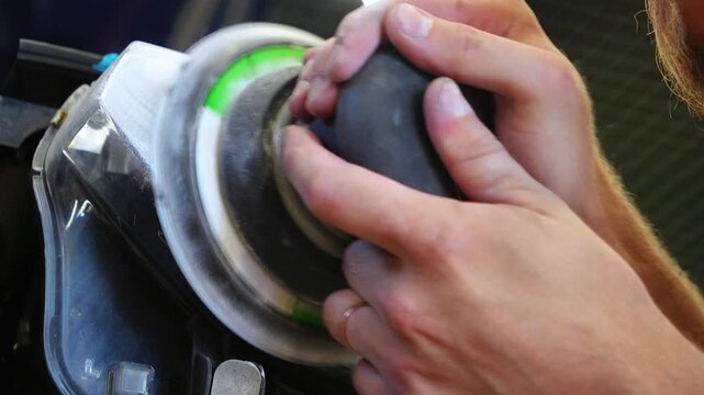 Car detailing technician sanding foggy plastic headlight close up, man hands using orbital sander machine with sandpaper for automotive headlamp restoration and scratch repair. 