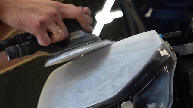 Car detailing technician sanding foggy plastic headlight close up, man hands using orbital sander machine with sandpaper for automotive headlamp restoration and scratch repair. 