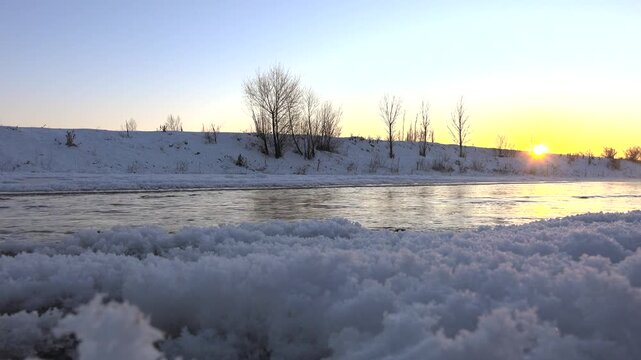 Sunrise lights snowy Canadian river as clear winter morning reveals flowing water and ice. Cold stream runs between frozen banks beneath cloudless dawn across North America.