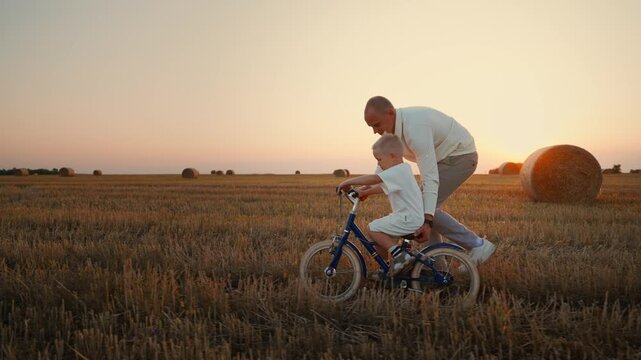 Father teaching young son to ride a bicycle through a golden harvested field at sunset, sharing a peaceful outdoor moment and creating warm family memories