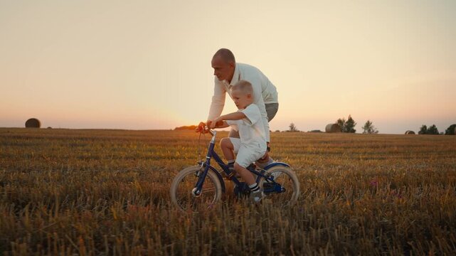 Father teaching young son to ride bicycle together in golden field at sunset, bonding over first cycling lesson and enjoying peaceful rural evening landscape