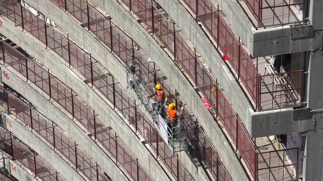 Construction workers on suspended platform repair skyscraper facade with insulation tools urban. Hanging scaffold team performs exterior building work, high rise maintenance scene industrial labor.