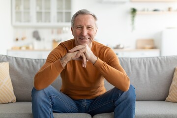 Fototapeta premium Smiling Mature Caucasian Man Seated Cross-legged On Couch In Cozy Living Room, Exuding Happiness And Contentment, Engaging In Relaxation And Enjoyment At Home. Family Lifestyle Concept.