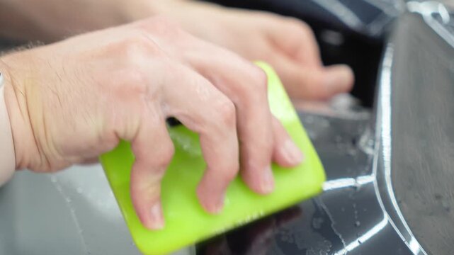 Professional auto detailer specialist applying a clear paint protection film PPF on a glossy black car. Male technician worker smoothing a wet polyurethane protective wrap with a green squeegee rakel.