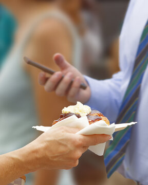 A woman holds an appetizing pastry topped with fruit compote and cream at the busy Naplavka farmers market in Prague, while a man in business attire checks his phone nearby.