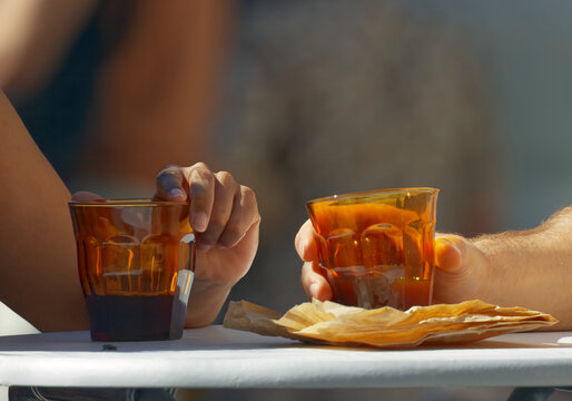 Two hands holding glasses of amber coffee backlit by warm sunlight, a crumpled kraft paper wrapper resting between them at the small, community-minded Hermanak farmers market in Prague.
