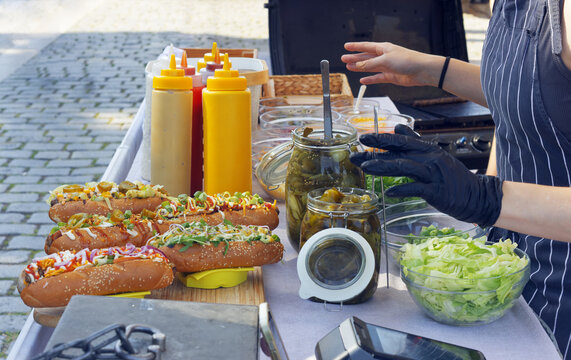 A gloved vendor prepares gourmet hot dogs beside condiment bottles, pickle jars and fresh lettuce on a cobblestone street at the Naplavka market in Prague.
