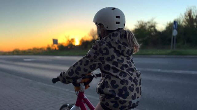 Helmeted child cycles steadily beside multi lane road as sun lowers behind trees. Small bicycle, patterned outerwear, warm evening glow, and distant traffic create active outdoor commute.
