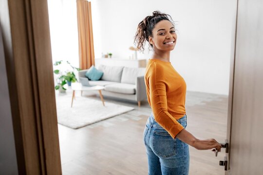 Joyful Smiling Black Woman Opening Door With Happiness, Standing In Modern Living Room, Welcoming Guests, In Cozy Home Environment. Home Interior Concept.