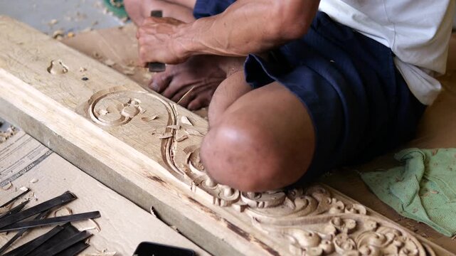 Wood carving continues on ornate panel, with man kneeling close and driving chisel into raised pattern. Curved detail, loose shavings, and scattered tools emphasize careful manual craft and precision.