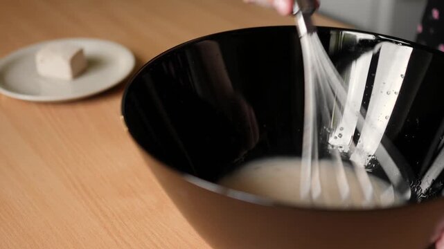 Close up side view of whisking yeast liquid mixture with wire balloon whisk in black bowl for dough preparation
