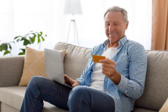 Smiling Senior Caucasian Man Using Laptop While Holding Credit Card For Online Shopping At Comfortable Living Room. Enjoying Convenience Of Digital Payment Options At Home. Business Concept.