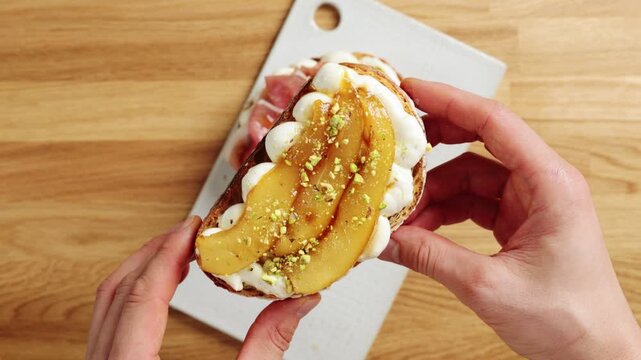 Hands show pear ricotta toast with nuts to camera. Seasonal cafe menu pairs creamy cheese with caramelized fruit. Elegant brunch dish suits coffee shop, bakery or autumn restaurant promotion.