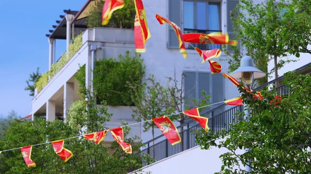 A string of small red Montenegro flags flutters in the breeze above a white residential building with balconies and green trees, a national pride decoration for a holiday or celebration.