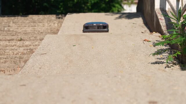 A small dark blue robot vacuum sits motionless on a concrete pathway at the top of stone steps, with greenery and a river visible in the background.