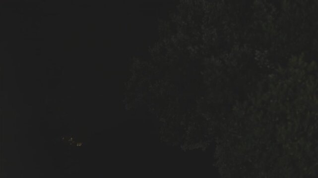 Flashes of lightning illuminate a dark night rain storm over a distant rural house surrounded by forest trees