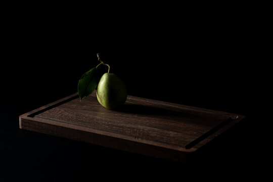 A single green pear with its stem and leaf attached rests on a dark wooden board against a black background, illuminated by dramatic side lighting, with generous negative space 
