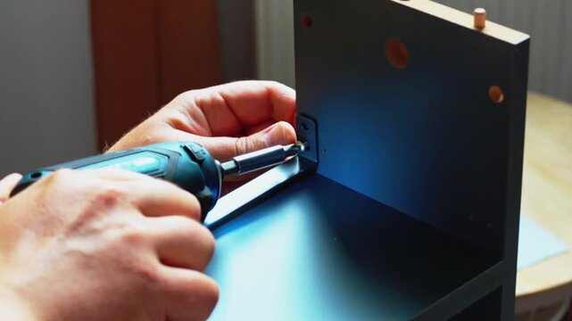 Close-up of male hands using a power screwdriver to assemble furniture, attaching a metal bracket to a wooden panel in a well-lit indoor setting