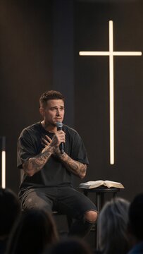 Man with tattoos preaching with microphone on stage with glowing cross and open bible. Young preacher sharing testimony at church service.