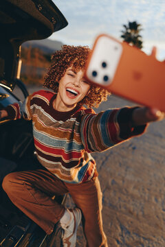 Woman selfie smile curly hair outdoors car lifestyle, young adult cheerful expression, striped sweater, leaning near open vehicle door, warm sunlight, urban summer travel relaxed mood