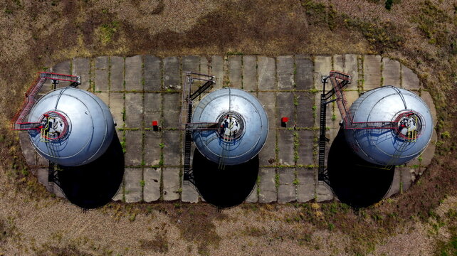 Aerial view of three large industrial spherical gas tanks situated on a concrete platform surrounded by dry grass in Belgrade, City of Belgrade, Serbia.