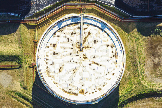 Aerial view of a large circular industrial storage tank with a central bridge structure, surrounded by grassy terrain and pipelines in La Spezia, Liguria, Italy.
