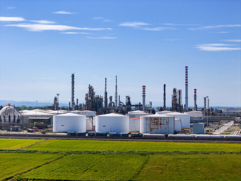 Aerial view of the Eni oil refinery with its large white storage tanks, industrial towers, and pipelines set against green fields in Sannazzaro de' Burgondi, Lombardy, Italy.