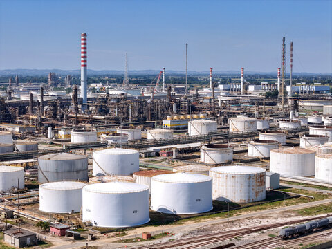 Aerial view of the Eni oil refinery featuring numerous large white storage tanks, industrial processing units, tall chimneys, and rail infrastructure in Sannazzaro de' Burgondi, Lombardy, Italy.
