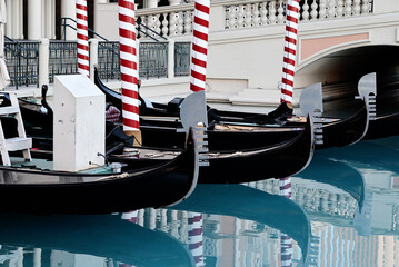 Several black gondolas are moored in a blue canal next to red-and-white striped posts. © Stuart