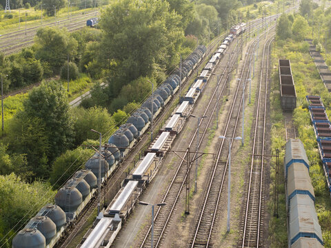 Aerial view of freight train with specialized tank wagons and flatbed cars carrying steel slabs on tracks through a green landscape in Poland.