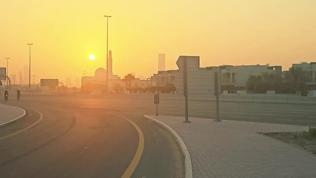 Golden sunrise over an urban street and paved path with mosque in hazy sunlight, creating a warm, peaceful, ambient morning landscape scene. People cycling on the road