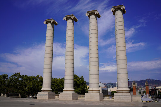 Four tall classical columns with Ionic capitals in Barcelona against a deep blue sky and scattered clouds