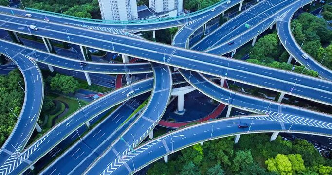 High angle shot of a complex multi-level highway interchange with sweeping curved overpasses, road markings, and green trees under cool daylight.