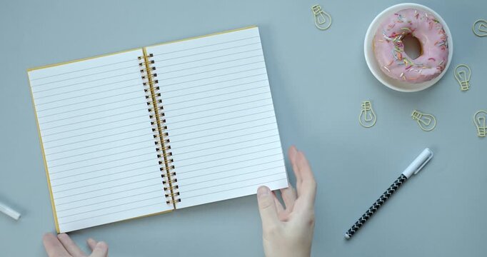 Hands turning pages of a notebook next to pink glazed donut and lightbulb clips stationery planner office paper spiral