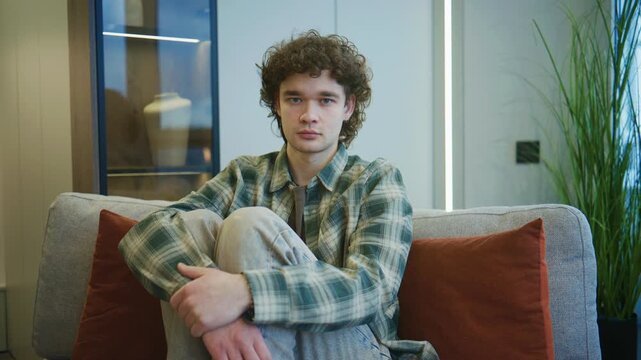 A young man sits on a sofa, hugging his knees to his chest, looking directly at the camera with a sad expression in a quiet home setting