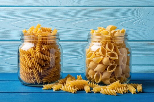 Two glass jars hold dry fusilli and conchiglie pasta. Uncooked pasta fills containers on blue wooden table. Ingredients ready for Italian meal preparation.