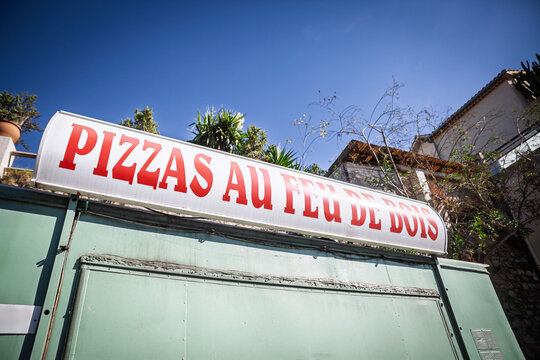 A large sign reading PIZZAS AU FEU DE BOIS, meaning wood fired pizza in french advertises above a roadside food stand, on a common French takeaway and pizzeria offering under a clear blue sky.