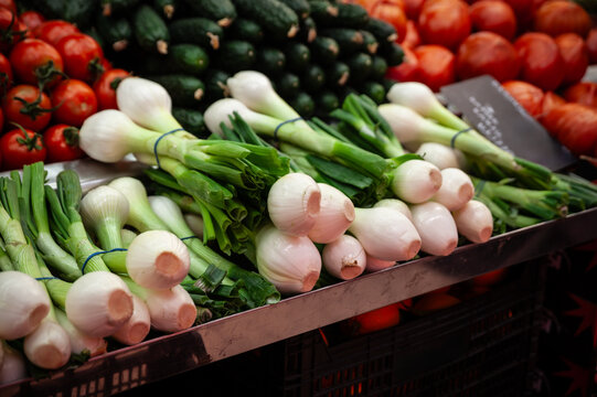 Close-up of bunches of young onions, also known as green onions or scallions, displayed for sale on a market stall, with tomatoes and cucumbers visible in the background.