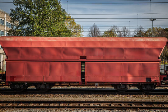 A red European hopper wagon for transporting bulk materials such as gravel and minerals, standing on railway tracks beneath overhead power lines, with trees and a building in the background.