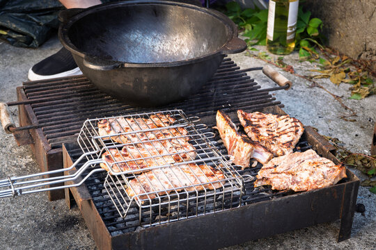 Pork chops cook over a charcoal barbecue, with browned grill marks visible on the meat and a large black cooking pot positioned above the rustic outdoor grill during open-air food preparation.
