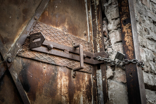 Close-up of a heavy rusty metal latch secured with a chain and padlock on an old weathered door, showing corrosion, aged hardware and rough masonry around a locked and deteriorating entrance.