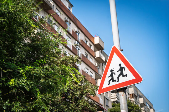 European children crossing warning sign on a roadside pole, with a red triangular border, bright blue sky, leafy trees and a softly blurred residential apartment building in an urban setting.