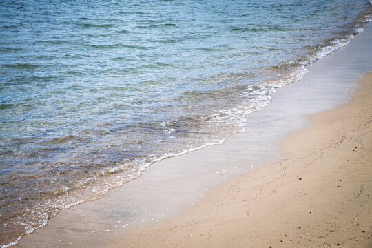 Close view of gentle Mediterranean Sea waves washing onto a sandy French beach, showing clear blue-green water, wet shoreline, foam and natural coastal textures along the edge of the sea.