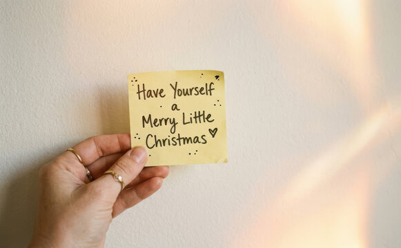 Hand holding yellow sticky note with Have Yourself a Merry Little Christmas quote message text handwritten greeting for holiday seasonal greeting card concept