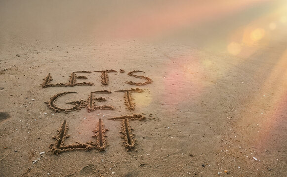 Let us get lit phrase written in sand on beach with warm sunlight flare representing summer party celebration and vacation mood