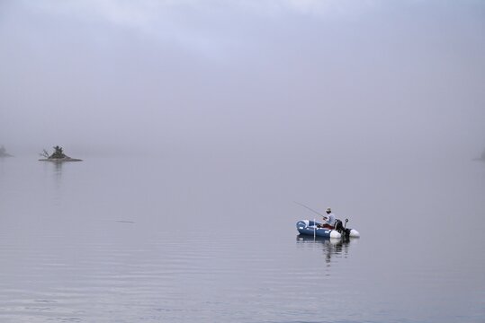 Small boat floating on a calm lake surrounded by dense fog, with a distant island fading into the mist. Atmospheric minimalist landscape with water and sky blending together, creating a serene and mys