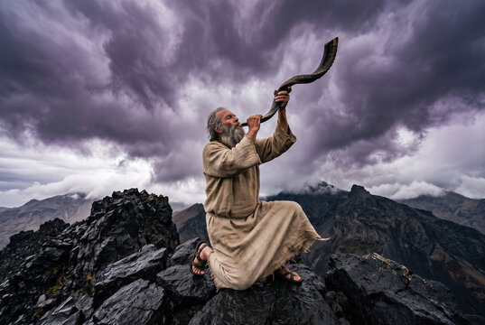 Ancient biblical prophet man kneeling on storm mountain blowing shofar horn in prayer.